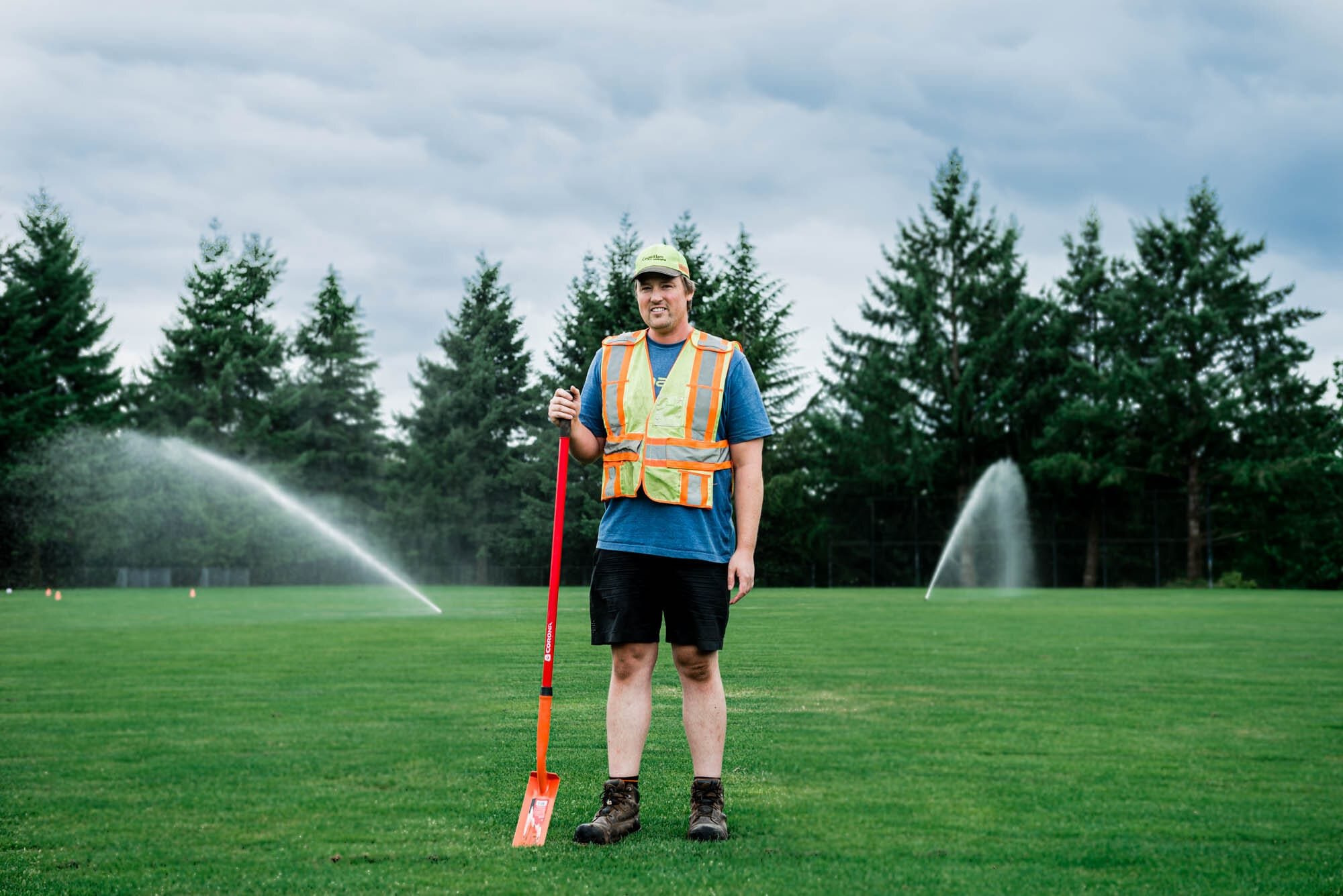 CoquitlamCityHR-5 City Irrigation Worker with shovel on grassy field with sprinklers.
